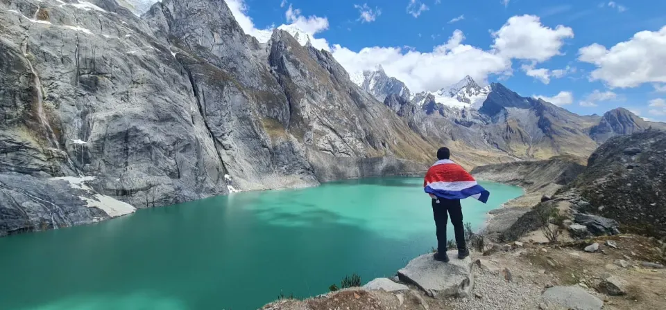 A trekker holding a Peruvian flag beside a turquoise glacial lake