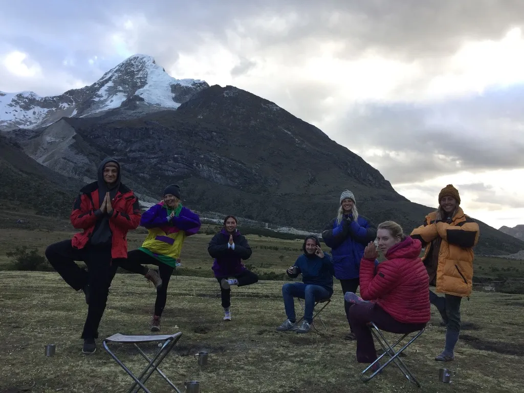 Trekkers in warm jackets posing beside camp stools beneath a snow-covered mountain
