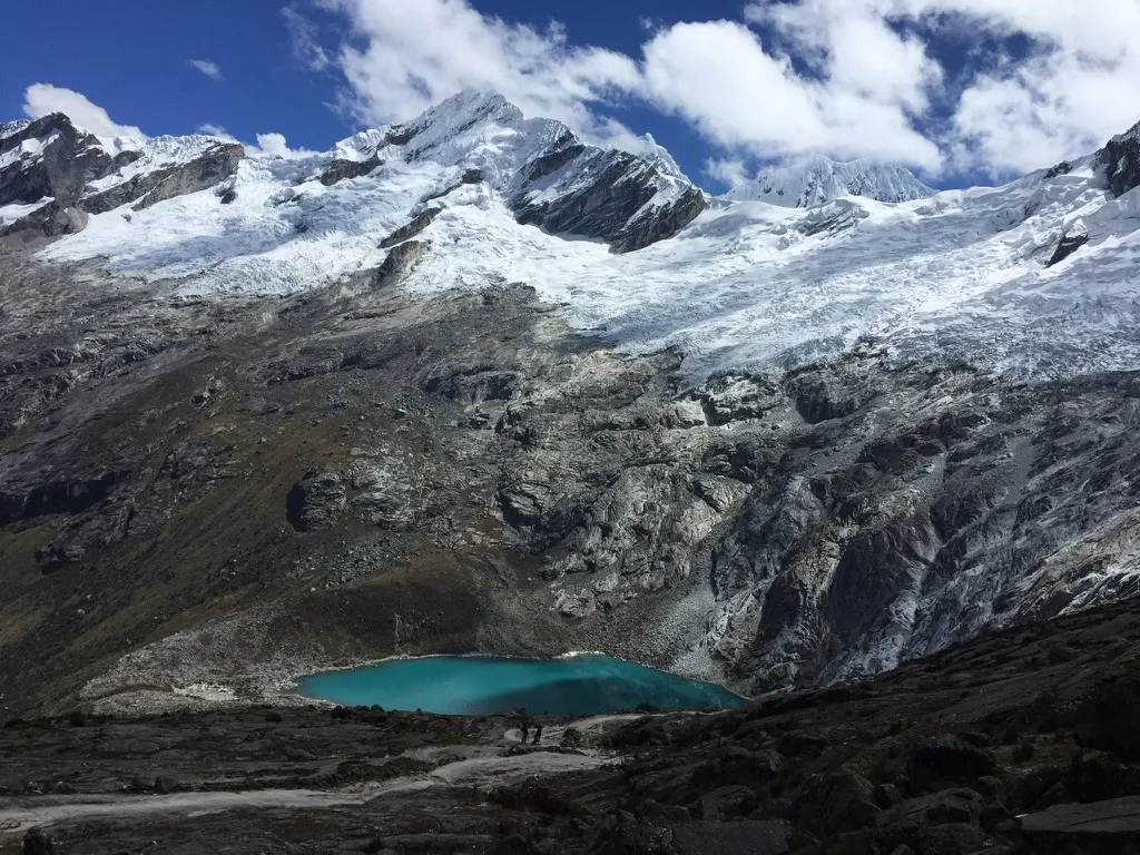 Trekkers on a rocky trail above a turquoise lake beneath glaciers and snow-covered peaks